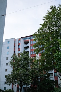 A multistory residential building with a modern design surrounded by green trees. The building has a white and red color scheme with multiple windows and balconies. The sky is slightly overcast, adding to the serene atmosphere.