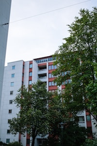 A multistory residential building with a modern design surrounded by green trees. The building has a white and red color scheme with multiple windows and balconies. The sky is slightly overcast, adding to the serene atmosphere.