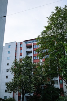 A multistory residential building with a modern design surrounded by green trees. The building has a white and red color scheme with multiple windows and balconies. The sky is slightly overcast, adding to the serene atmosphere.