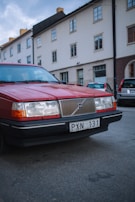 A red Volvo car is parked on the side of a quiet residential street. The car is an older model, characterized by its boxy shape and distinctive front grille. In the background, there are several buildings with white and brown exteriors, and another car is parked behind the Volvo.