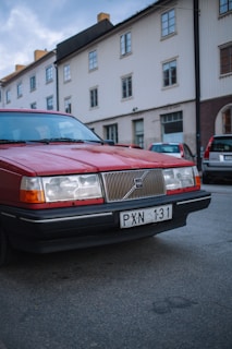 A red Volvo car is parked on the side of a quiet residential street. The car is an older model, characterized by its boxy shape and distinctive front grille. In the background, there are several buildings with white and brown exteriors, and another car is parked behind the Volvo.