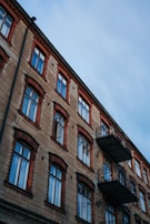 A multi-story residential building with a classic architectural design features rows of windows and two protruding metal balconies. The building facade is made of stone with contrasting red brick accents surrounding each window, providing a vintage aesthetic. The sky above is overcast with a cool, bluish hue.
