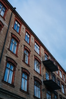 A multi-story residential building with a classic architectural design features rows of windows and two protruding metal balconies. The building facade is made of stone with contrasting red brick accents surrounding each window, providing a vintage aesthetic. The sky above is overcast with a cool, bluish hue.