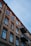A multi-story residential building with a classic architectural design features rows of windows and two protruding metal balconies. The building facade is made of stone with contrasting red brick accents surrounding each window, providing a vintage aesthetic. The sky above is overcast with a cool, bluish hue.