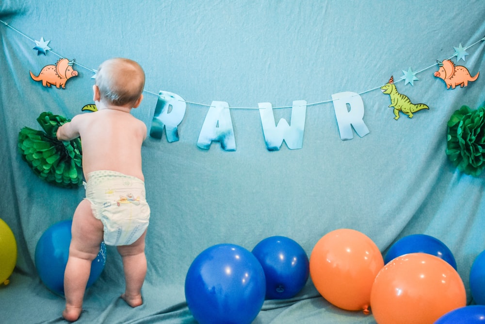 baby in white diaper standing beside blue and red balloons