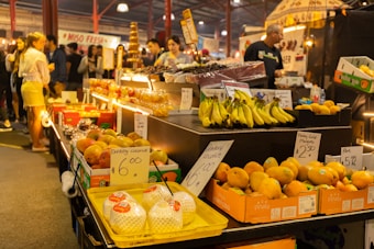 An indoor market scene with various fruits and food items displayed on a counter. Boxes of bananas, mangoes, and drinking coconuts are prominently visible. Price tags are displayed for many items, such as drinking coconuts for $6 and honey gold mangoes for $2.50 each. There are people in the background, some appearing to be customers and others possibly vendors. The atmosphere is busy and vibrant, with warm lighting giving a cozy feel.