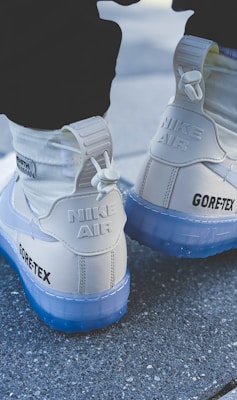 A pair of white Nike Air shoes with Gore-Tex branding is shown from the back. The shoes have a chunky sole with a translucent blue tint and a high-top design. They are being worn with white socks, and the surface beneath is a textured gray pavement.