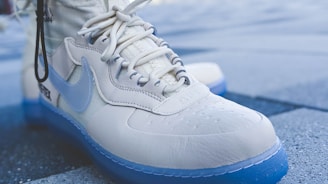 A close-up of a sneaker with a prominent glossy white upper material and a translucent blue sole. The shoe features detailed stitching, a textured surface, and a lace-up design. The well-known brand logo is visible on the side.