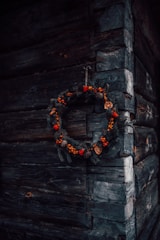 A rustic wooden wall corner with a decorative wreath hanging, made of pine branches, red berries, pine cones, and dried orange slices.