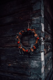 A rustic wooden wall corner with a decorative wreath hanging, made of pine branches, red berries, pine cones, and dried orange slices.