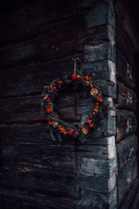 A rustic wooden wall corner with a decorative wreath hanging, made of pine branches, red berries, pine cones, and dried orange slices.