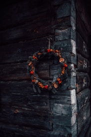 A rustic wooden wall corner with a decorative wreath hanging, made of pine branches, red berries, pine cones, and dried orange slices.