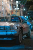 A futuristic car with a stainless steel body and prominent blue neon lighting is parked in an urban setting at night. The car features a distinctive design with a wide front grille, stylish wheel rims, and external modifications that enhance its sci-fi appearance. The background reveals a blurred urban landscape with additional lights and structures.