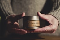 Two human hands hold a small, round container labeled 'Le Petit Raspberry Soy Wax Hand Poured'. The hands are wearing a knitted sweater. The lighting is warm, casting soft shadows, and the background is softly blurred.