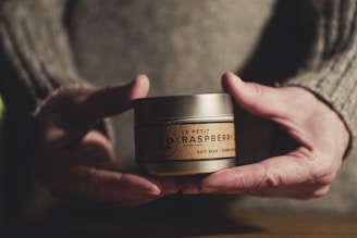 Two human hands hold a small, round container labeled 'Le Petit Raspberry Soy Wax Hand Poured'. The hands are wearing a knitted sweater. The lighting is warm, casting soft shadows, and the background is softly blurred.