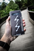 A hand holds a smartphone with a black case featuring a lightning bolt design and the text 'BEWARE: HIGH VOLTAGE'. The phone is being held over a wooden walkway with surrounding green foliage.