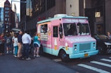 A brightly colored ice cream truck with a pink and blue exterior is parked on a city street. A group of people is gathered around, with some placing orders and others waiting. The side of the truck displays images of ice cream cones and shakes.