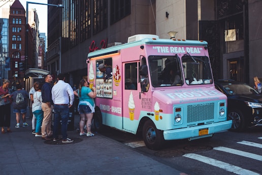 A brightly colored ice cream truck with a pink and blue exterior is parked on a city street. A group of people is gathered around, with some placing orders and others waiting. The side of the truck displays images of ice cream cones and shakes.