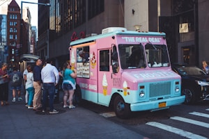 A brightly colored ice cream truck with a pink and blue exterior is parked on a city street. A group of people is gathered around, with some placing orders and others waiting. The side of the truck displays images of ice cream cones and shakes.