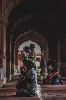 People are engaged in prayer within a dimly lit, intricately designed mosque interior. Their postures are reverent and solemn, some kneeling and others standing. The architecture features ornate arches and patterns, contributing to an atmosphere of tranquility and devotion.