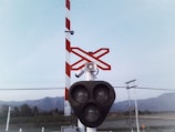 A railway crossing signal with three large circular lights is positioned under red and white striped barriers. A red and white crossbuck sign is visible above. There are telephone poles and power lines in the background with a view of mountains and trees under a pale blue sky.