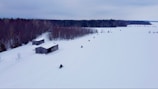 Several snowmobiles are moving across a vast snowy landscape with two wooden sheds nearby. A dense forest in the background contrasts with the white snowfield.