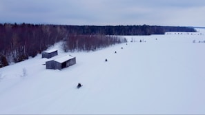 Several snowmobiles are moving across a vast snowy landscape with two wooden sheds nearby. A dense forest in the background contrasts with the white snowfield.