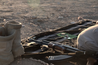 A rifle lies on the ground beside a pair of boots and some personal items, including a watch and a pair of glasses, on a piece of fabric over a rocky, sandy surface.
