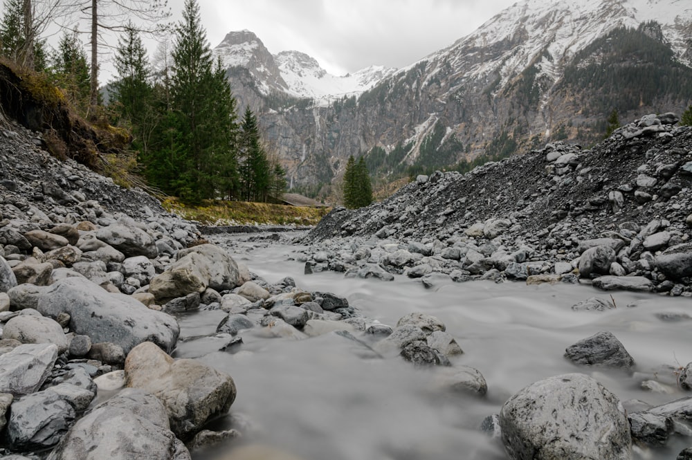 rocky river near green trees and snow covered mountain during daytime