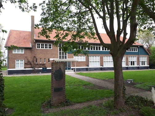 A large, two-story brick building with a red-tiled roof and multiple white-framed windows stands prominently. A tree in the foreground partially obscures the view, its leaves casting shadows on the lawn. A small brick structure with a plaque is situated near the building. The surrounding area is lush with green grass and a few scattered shrubs.