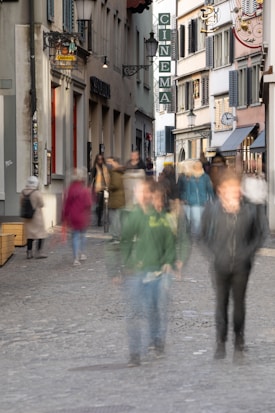 A cobblestone street lined with old buildings featuring a large cinema sign. The street is populated with blurred figures of people, capturing a sense of movement. Some storefronts and signs are visible, and a decorative wall clock is mounted on one building.