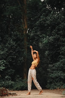 A person is standing on a path surrounded by dense green foliage, stretching or dancing with arms raised above the head. They are wearing a yellow crop top and white pants with an artistic pattern. The setting appears to be a forest or nature area with a serene and tranquil atmosphere.