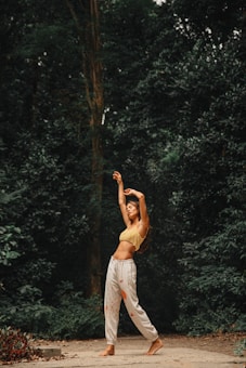A person is standing on a path surrounded by dense green foliage, stretching or dancing with arms raised above the head. They are wearing a yellow crop top and white pants with an artistic pattern. The setting appears to be a forest or nature area with a serene and tranquil atmosphere.