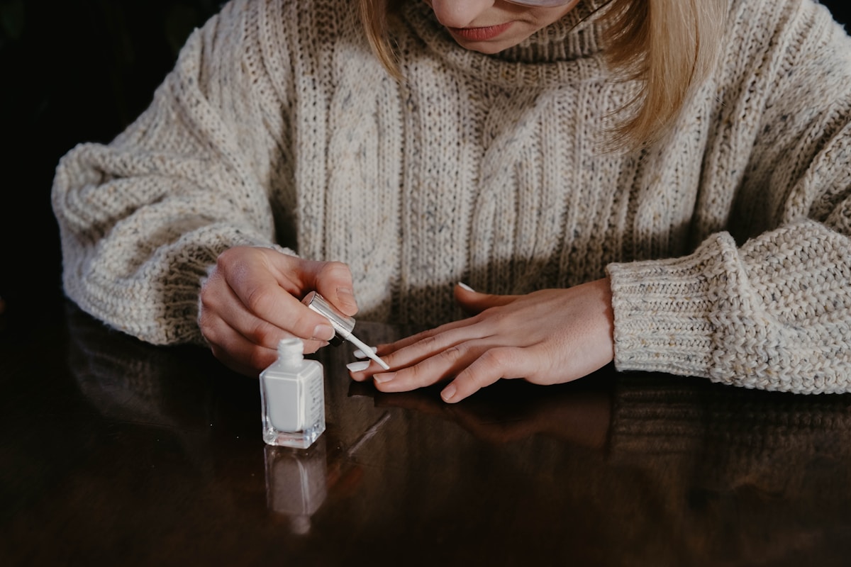 Woman in sweater painting her nails with white nail polish on table