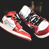 Pair of red, white, and black high-top sneakers with a prominent logo text on the side. The sneakers feature intricate designs with visible laces and are positioned creatively against a dark background.