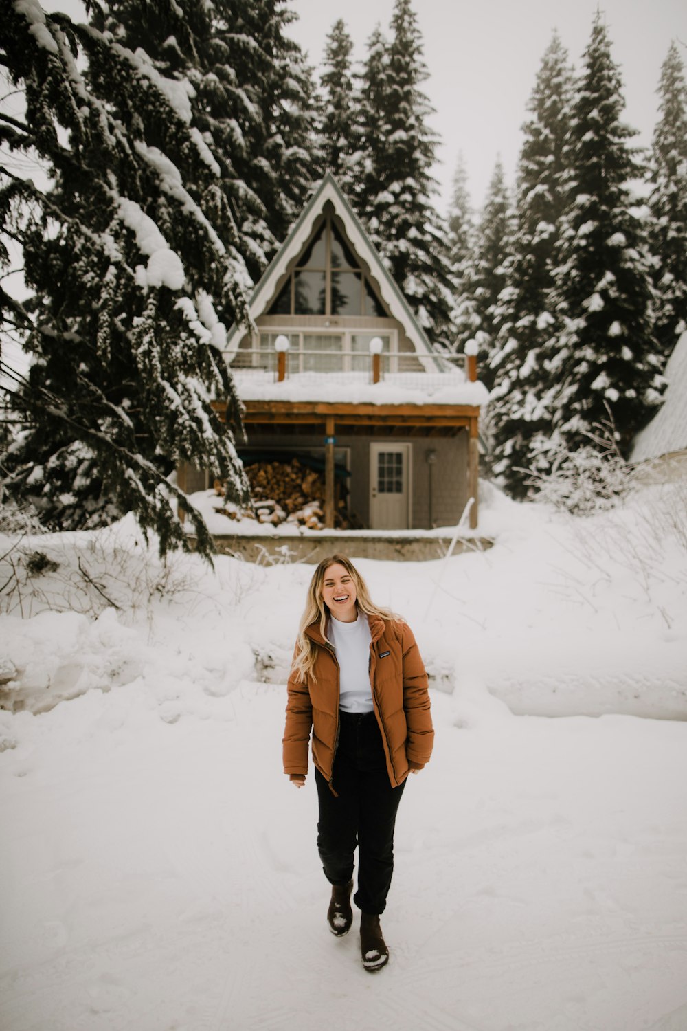 woman in brown coat standing on snow covered ground near brown wooden house during daytime