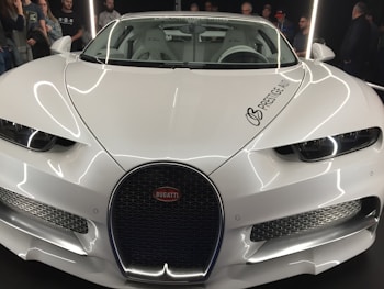 A sleek and elegant white Bugatti car is prominently displayed, surrounded by a group of people who appear to be admiring it. The brand logo and distinctive grille are visible, highlighting its luxury design. The setting suggests an indoor showroom with bright lighting reflecting off the car's polished surface.