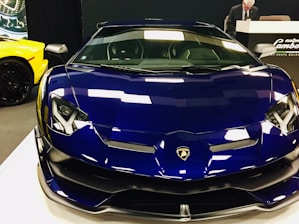A sleek blue Lamborghini sports car is displayed prominently in a showroom setting. The glossy surface reflects overhead lighting, emphasizing its angular design and luxurious appearance. In the background, part of a yellow car is visible, along with a person standing at a counter with branding signage.