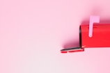 A red mailbox is positioned against a pastel pink background, with the mailbox door open and a pink flag raised.