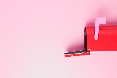 A red mailbox is positioned against a pastel pink background, with the mailbox door open and a pink flag raised.