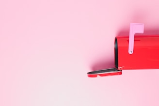 A red mailbox is positioned against a pastel pink background, with the mailbox door open and a pink flag raised.