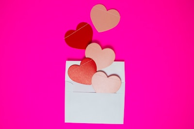 A white envelope lies open on a vibrant pink background. Several heart-shaped cutouts, in shades of red and pink, are placed inside the envelope and appear to be floating or spilling out.