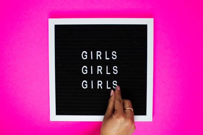 A black letter board with white letters spelling 'GIRLS' repeated three times, set against a bright pink background. A person's hand with neatly manicured nails is adjusting the letters on the board.