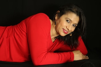 A woman with dark hair and a bright smile is lying down, wearing a vibrant red top. She is adorned with gold earrings against a black background, creating a stark contrast.