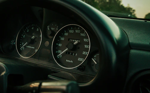 A close-up view of a car dashboard featuring a speedometer, tachometer, and other gauges. The dials display various units and measurements, such as kilometers per hour for speed and revolutions per minute for engine speed. The lighting is dim, contributing to a muted atmosphere.