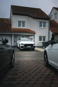 A modern, two-story house with a red-tiled roof and white exterior walls. A white car is parked in the driveway, surrounded by two other cars partially visible from the sides. The house features windows with white frames and a garage door.