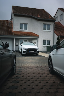 A modern, two-story house with a red-tiled roof and white exterior walls. A white car is parked in the driveway, surrounded by two other cars partially visible from the sides. The house features windows with white frames and a garage door.