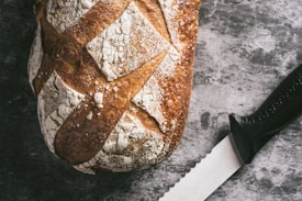 A rustic loaf of bread with a crispy, textured crust rests beside a serrated knife on a gray, textured surface. The bread features a pattern of flour dusting, highlighting its artisanal nature.