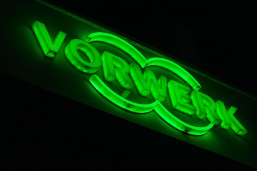 A neon green illuminated sign displaying the word 'VORWERK' with a double oval design around the letters.