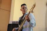 A man is playing an electric bass guitar while looking focused. He is wearing a light denim jacket over a black t-shirt. In the background, there is an amplifier, and the setting appears to be indoors, with textured cream-colored walls.